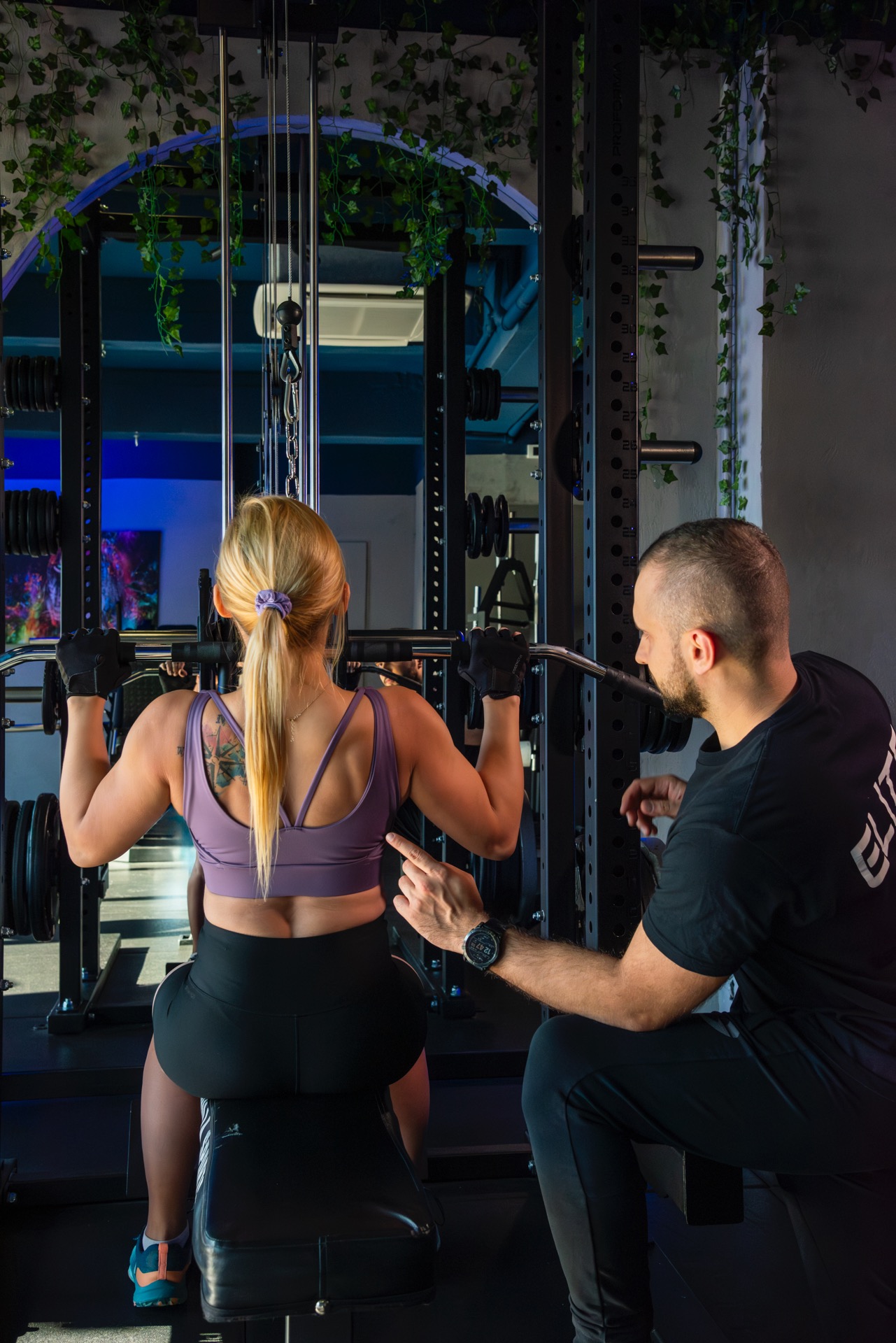 Kobieta ćwiczy na maszynie pod okiem trenera w siłowni. Instruktor wskazuje na technikę wykonywania ćwiczenia. W tle dekoracje z roślin.