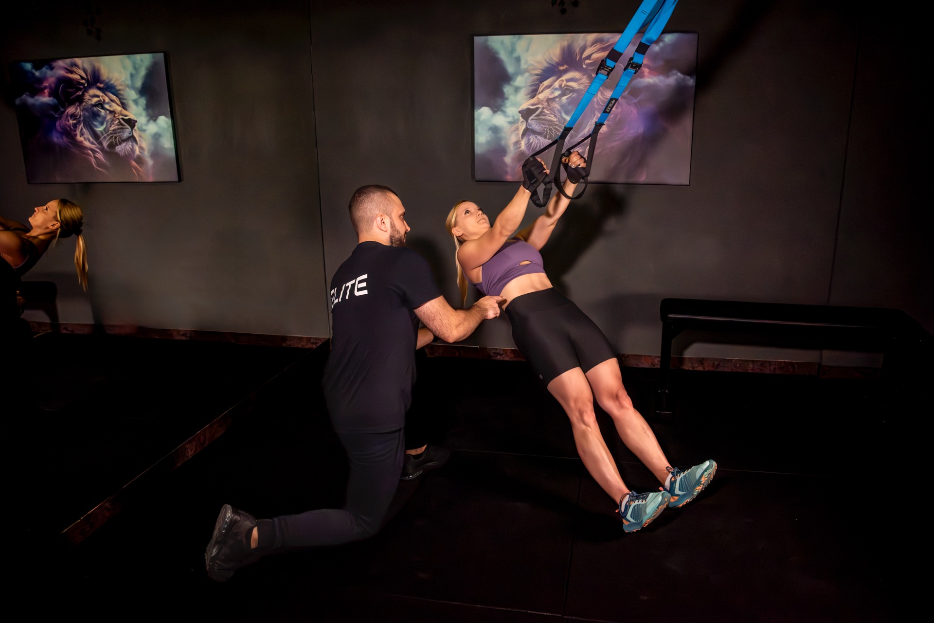 Trener asekuruje ćwiczącą kobietę podczas treningu na taśmach TRX w ciemnym pomieszczeniu z motywem lwa na ścianie.