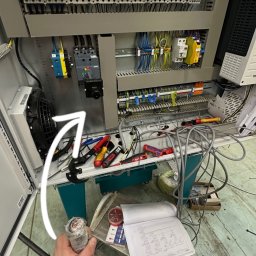 Konrad Hołubowicz Synergy - Panel elektryczny w trakcie prac: widoczne kable, narzędzia i schemat. Ręka elektryka trzyma kabel. Elementy instalacji w szarej obudowie.