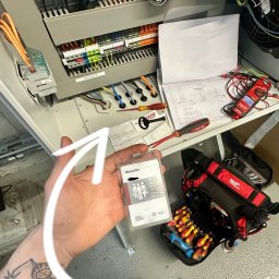 Konrad Hołubowicz Synergy - Otwarta szafa sterownicza z widocznymi bezpiecznikami, okablowaniem i schematem. W tle narzędzia, w ręku identyfikator BMW Group. Widoczny tatuaż na ręce.