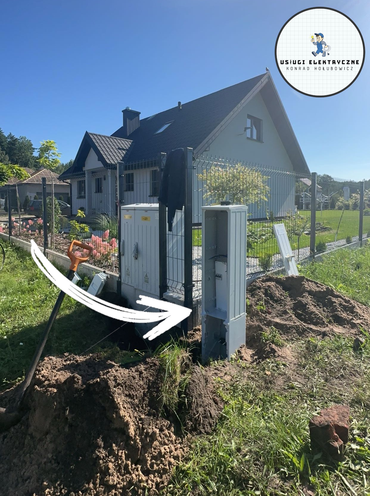 Przyłącze elektryczne do domu w trakcie realizacji: skrzynka ZK (złącze kablowe) i rozdzielnia główna na tle domu jednorodzinnego i ogrodzenia. Widoczna łopata i wykop.