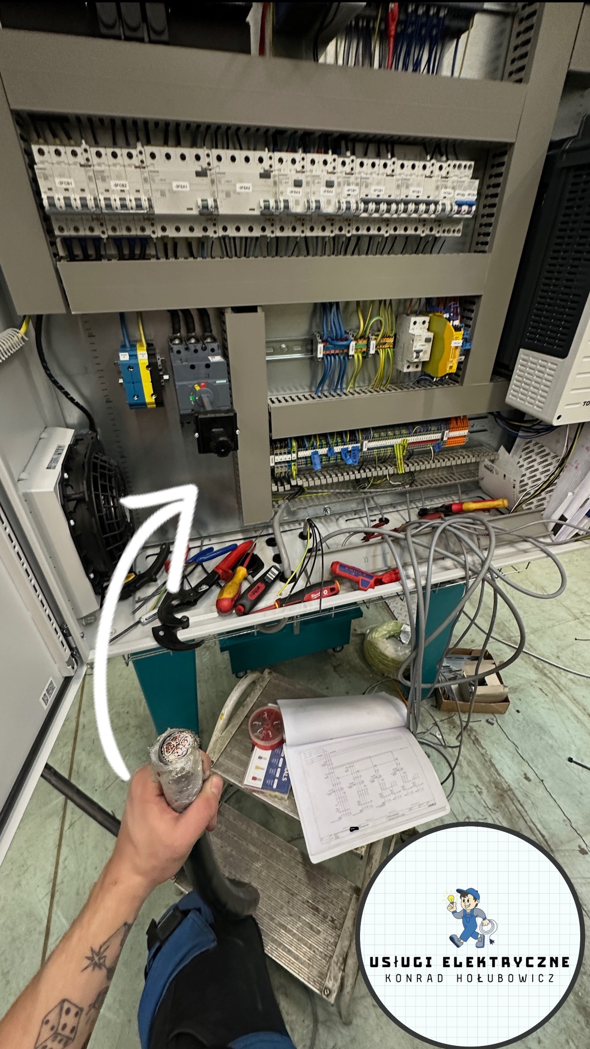 Panel elektryczny w trakcie prac: widoczne kable, narzędzia i schemat. Ręka elektryka trzyma kabel. Elementy instalacji w szarej obudowie.