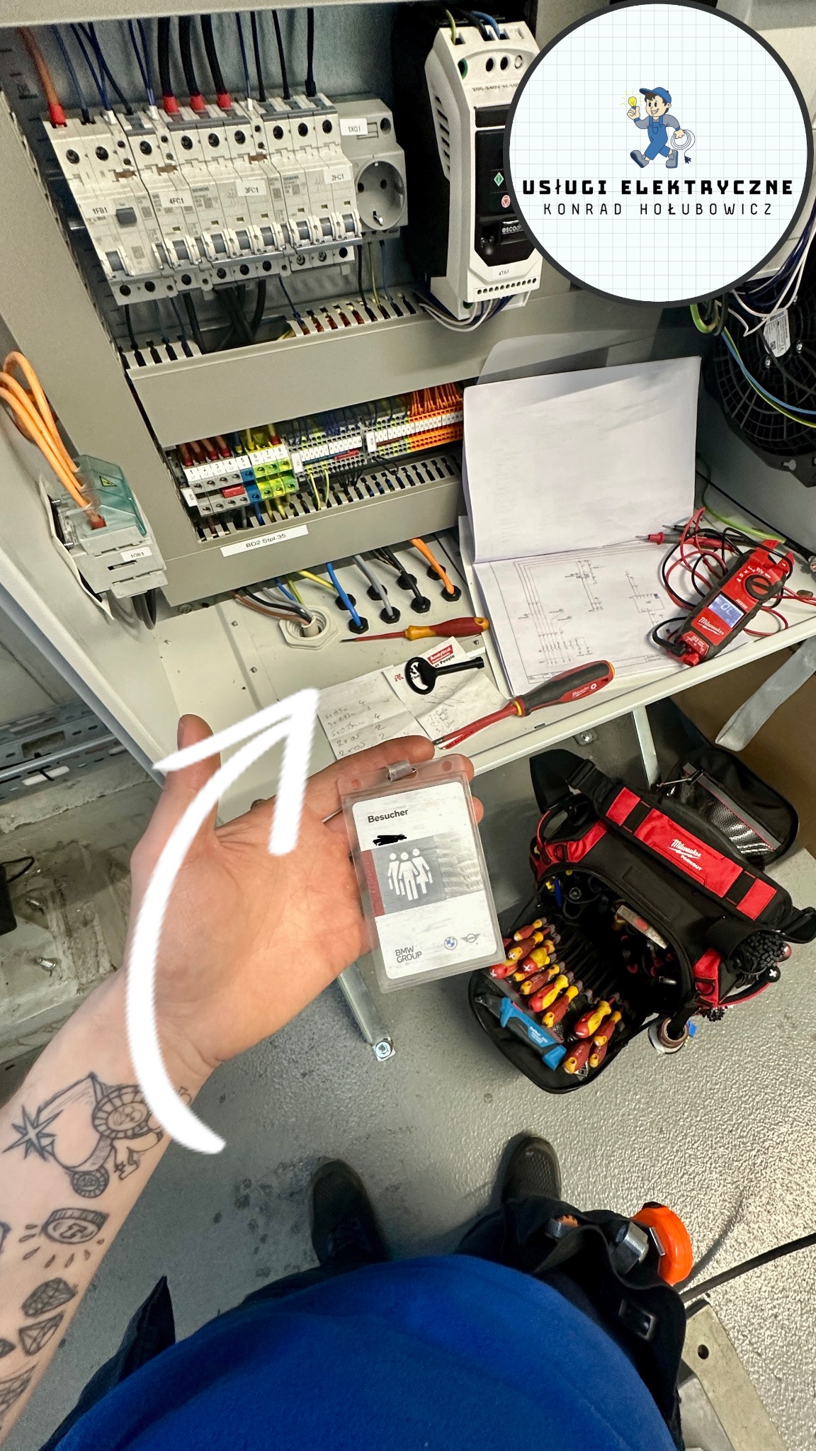 Otwarta szafa sterownicza z widocznymi bezpiecznikami, okablowaniem i schematem. W tle narzędzia, w ręku identyfikator BMW Group. Widoczny tatuaż na ręce.