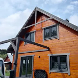 Drewniany dom z ciemnymi oknami i elementami wykończeniowymi, widziany z perspektywy niskiego kąta, z fragmentem białego samochodu z pomarańczowym kogutem.