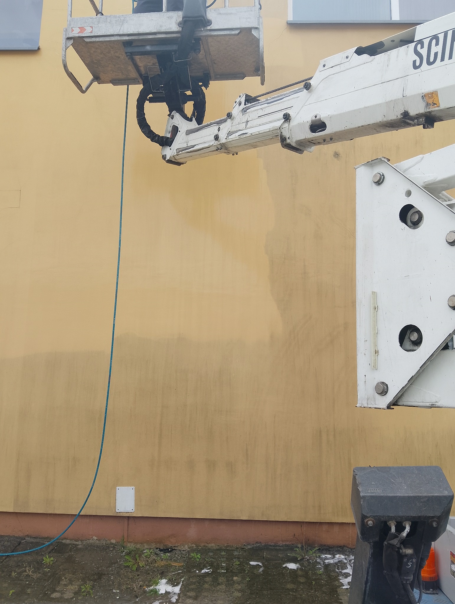 Żółta elewacja budynku w trakcie mycia pod ciśnieniem z podnośnika koszowego. Widoczne ślady czyszczenia. Część elewacji jest już umyta, a część jeszcze zabrudzona.