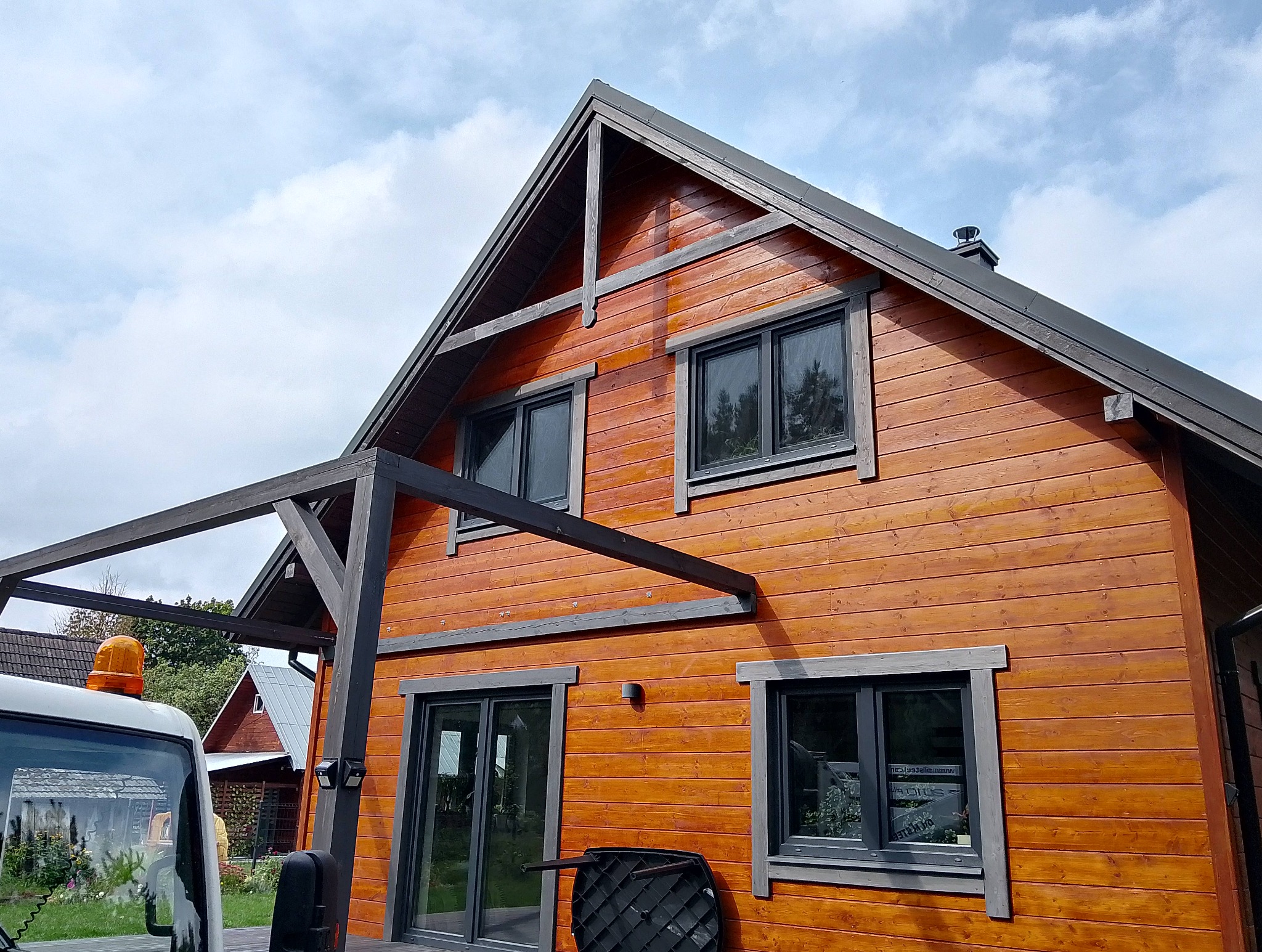 Drewniany dom z ciemnymi oknami i elementami wykończeniowymi, widziany z perspektywy niskiego kąta, z fragmentem białego samochodu z pomarańczowym kogutem.