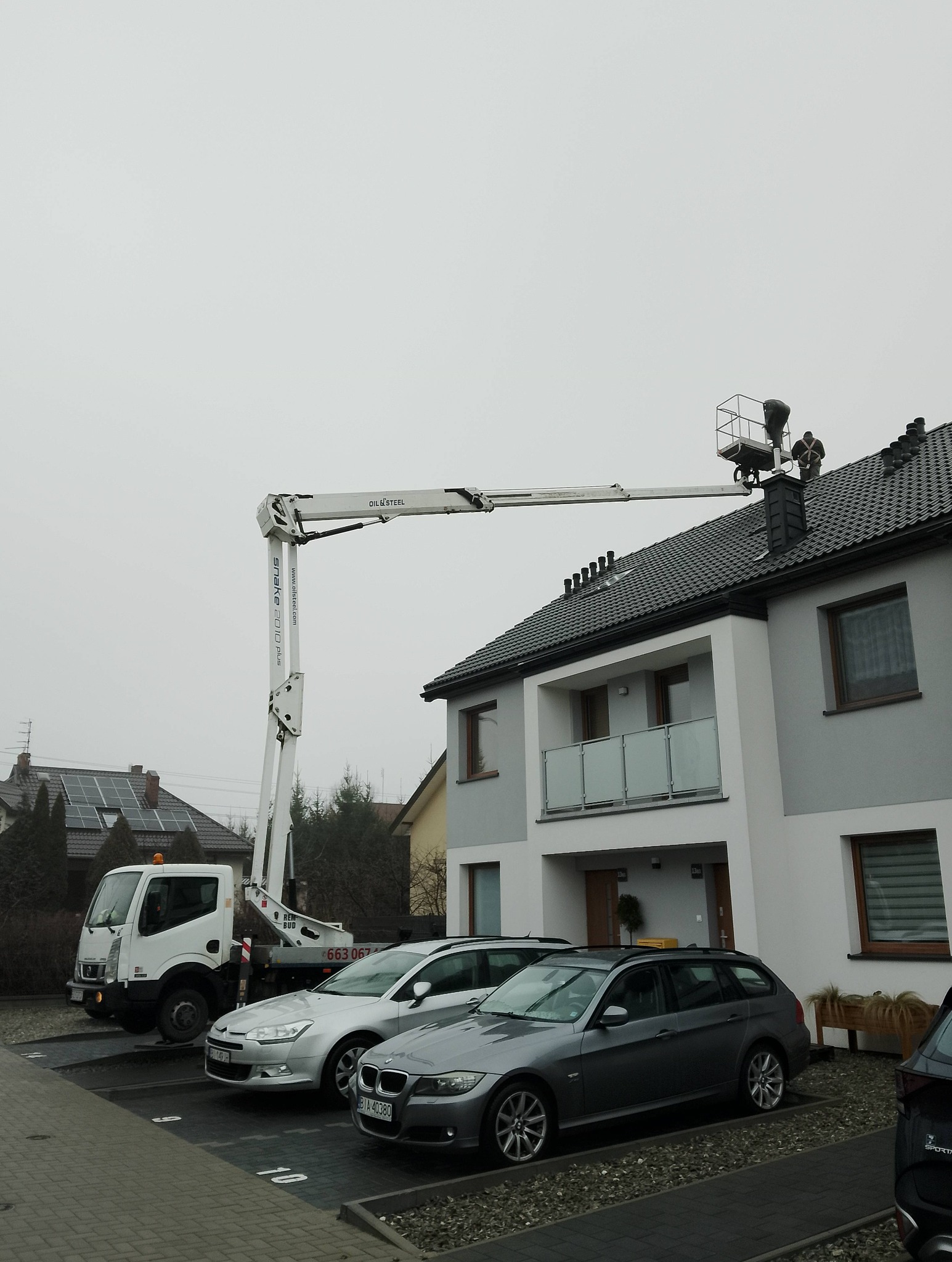 Pracownicy na dachu domu jednorodzinnego, pracujący z podnośnika koszowego. Widoczny fragment elewacji i zaparkowane samochody. Pochmurne niebo.