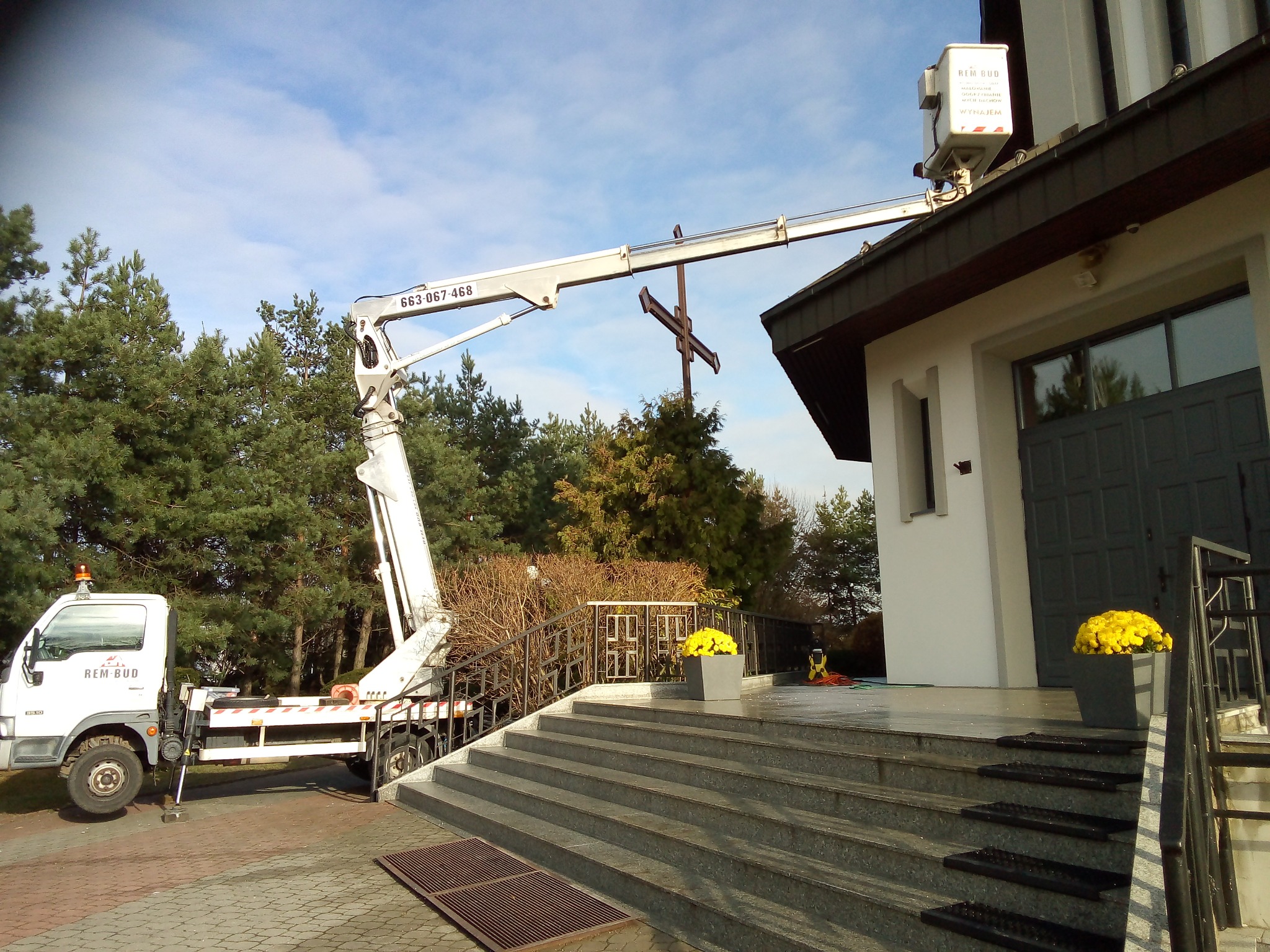 Podnośnik koszowy firmy REM-BUD przy kościele, ustawiony do prac na wysokości, z krzyżem w tle. Kwiaty w donicach zdobią wejście.
