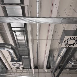 Tilda AP - Szara instalacja wentylacyjna pod sufitem, widoczne rury, kanały i kratki wentylacyjne. Na dole kadru drabina z pracownikiem w czarnym ubraniu.