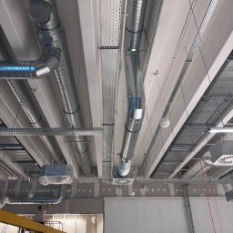Tilda AP - Montaż sufitowej instalacji wentylacyjnej z metalowych rur i kanałów w budynku komercyjnym. Widoczne elementy mocujące i kratki wentylacyjne. Prace w toku.