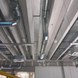 Montaż sufitowej instalacji wentylacyjnej z metalowych rur i kanałów w budynku komercyjnym. Widoczne elementy mocujące i kratki wentylacyjne. Prace w toku.
