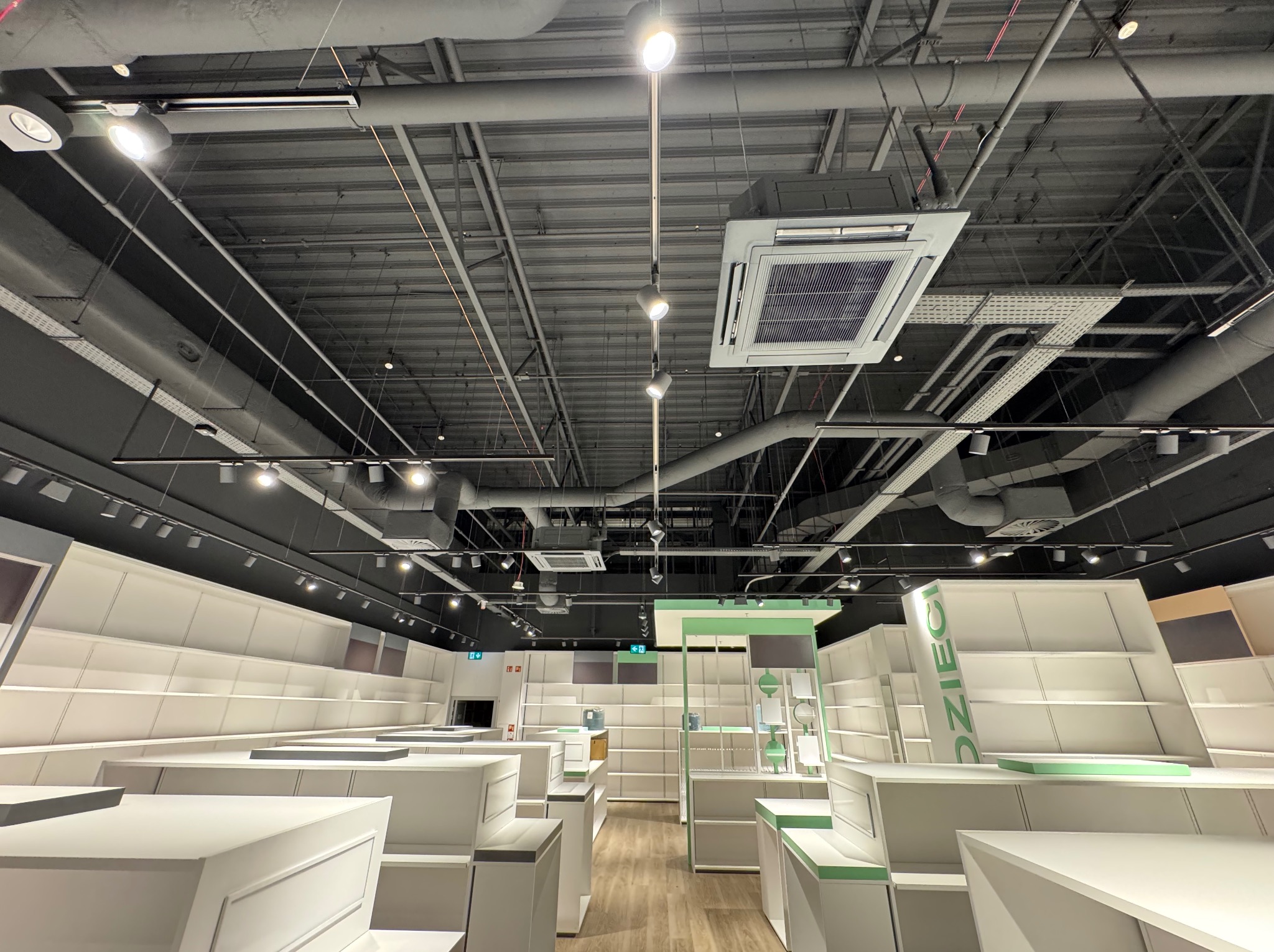 Sufit w sklepie z widocznymi instalacjami wentylacyjnymi i klimatyzacyjnymi, oświetleniem oraz elementami konstrukcyjnymi. Widoczne kanały i kratki wentylacyjne.