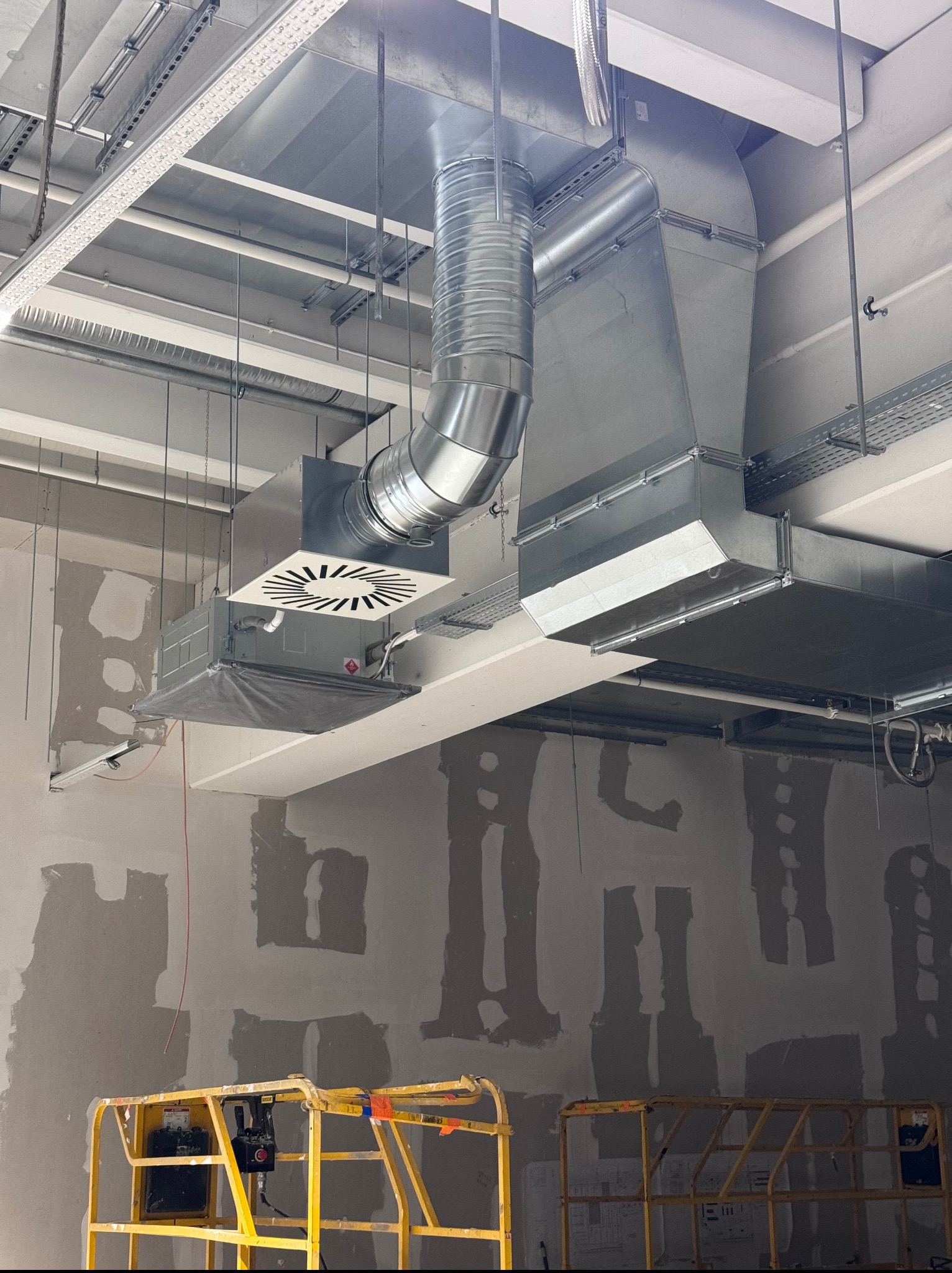Metalowe kanały wentylacyjne pod sufitem w surowym wnętrzu. Żółty podnośnik koszowy w dolnej części kadru. Widoczne elementy montażowe.