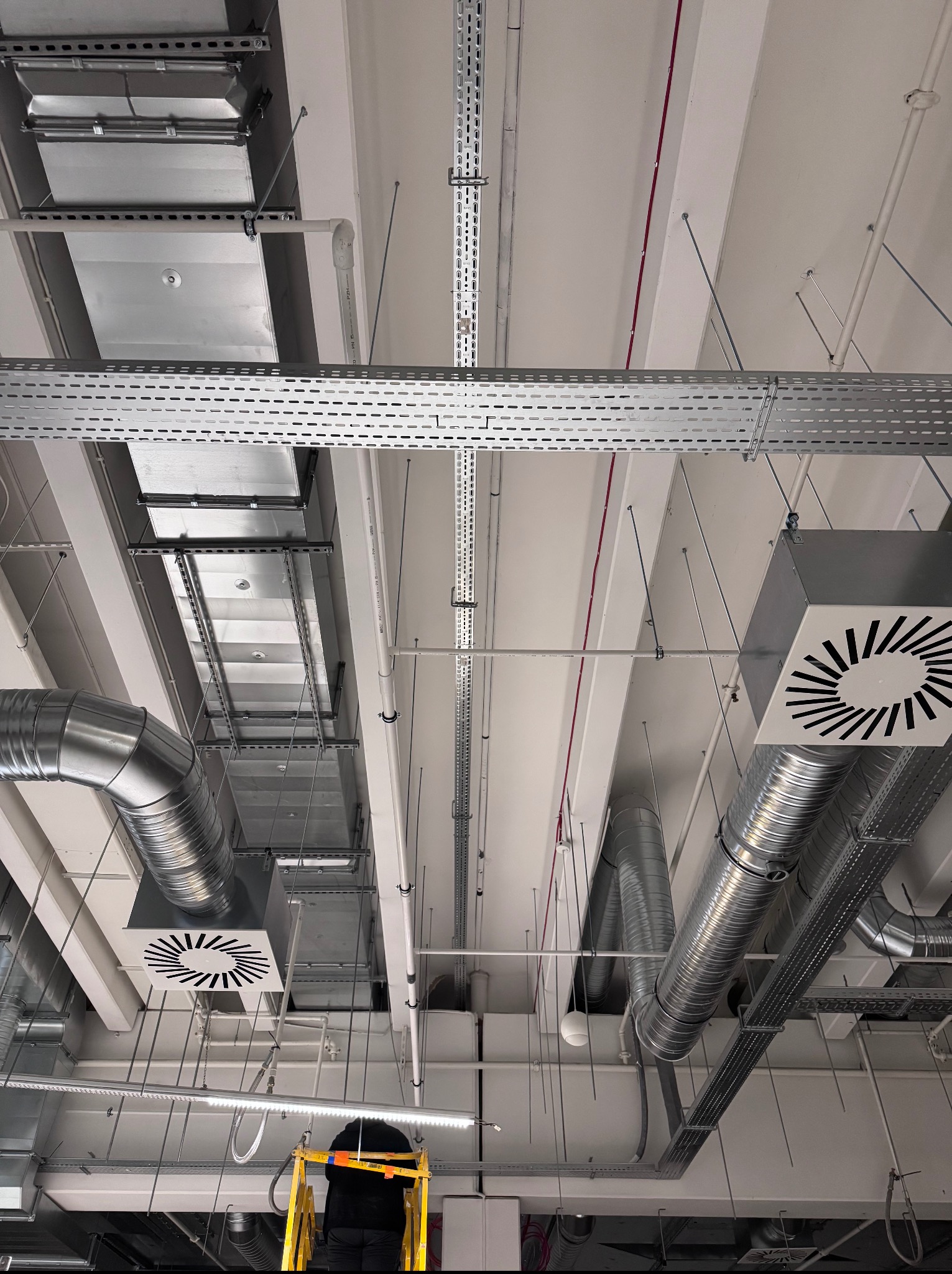 Szara instalacja wentylacyjna pod sufitem, widoczne rury, kanały i kratki wentylacyjne. Na dole kadru drabina z pracownikiem w czarnym ubraniu.