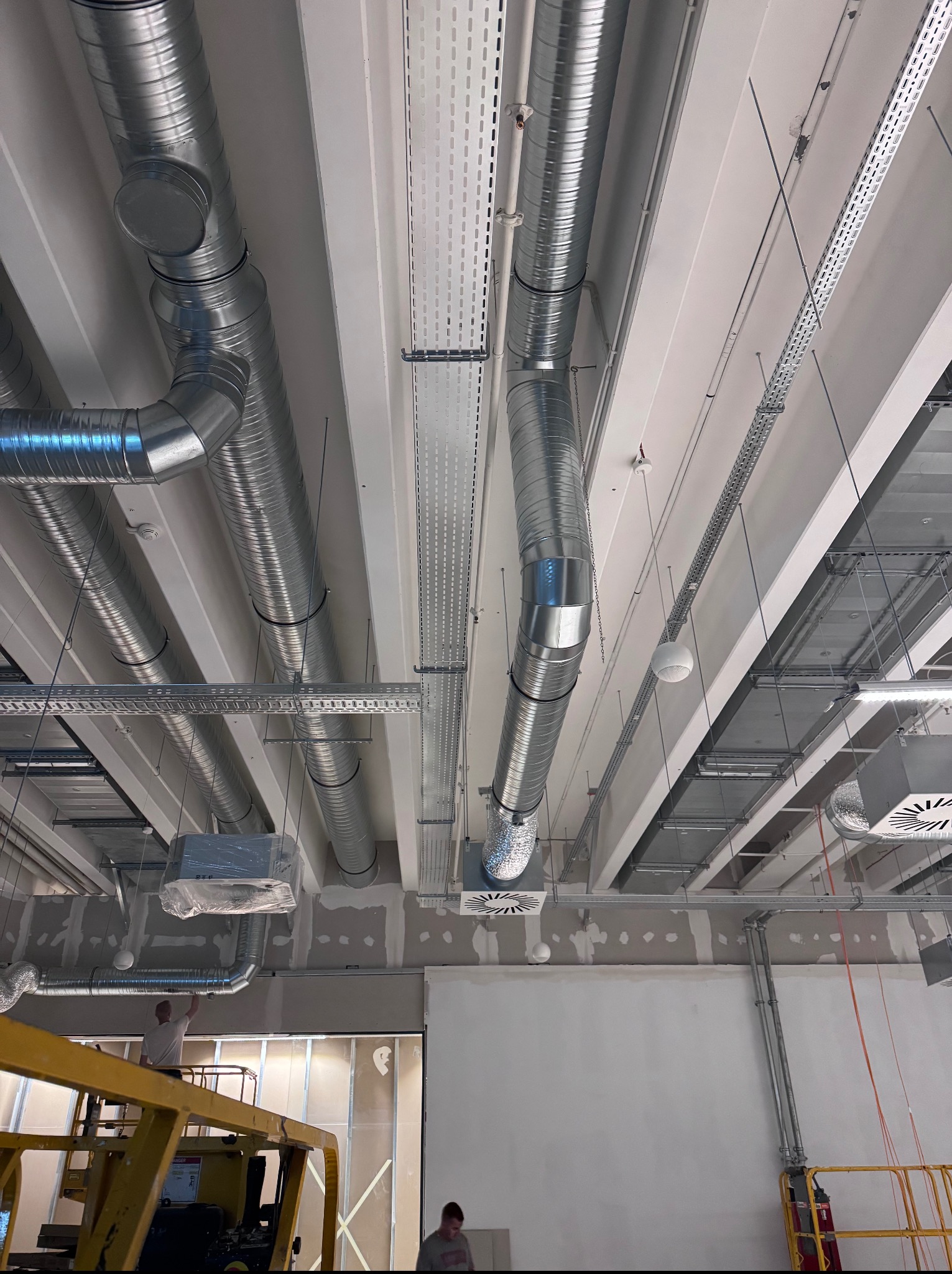 Montaż sufitowej instalacji wentylacyjnej z metalowych rur i kanałów w budynku komercyjnym. Widoczne elementy mocujące i kratki wentylacyjne. Prace w toku.