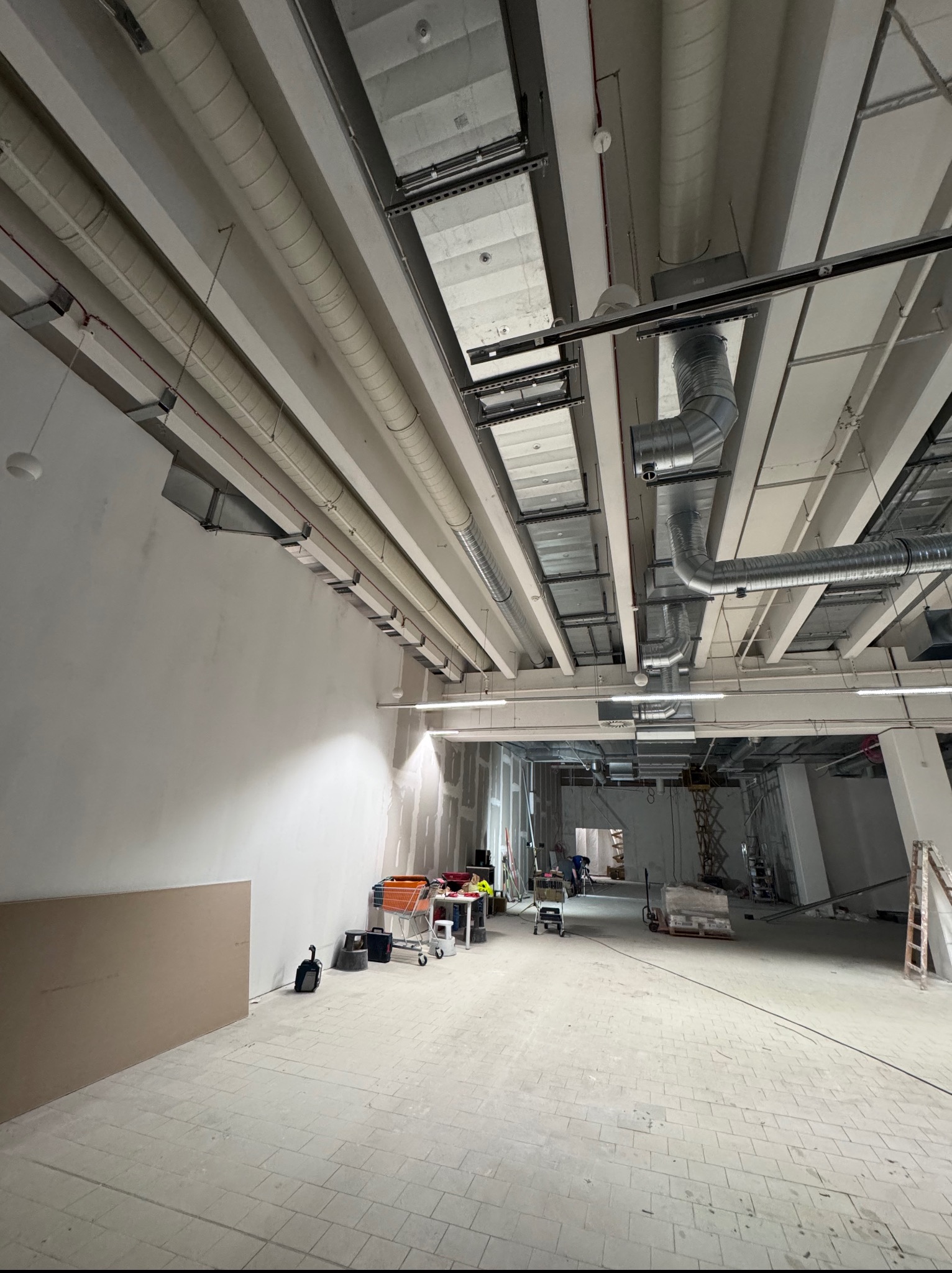 Wnętrze w trakcie instalacji wentylacji: widoczne rury wentylacyjne pod sufitem, narzędzia i materiały budowlane, surowy stan wykończenia pomieszczenia.