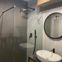 Nowoczesna łazienka z kabiną typu walk-in, owalną umywalką nablatową i okrągłym lustrem w czarnej ramie. Minimalistyczny design z elementami drewna i szarości.