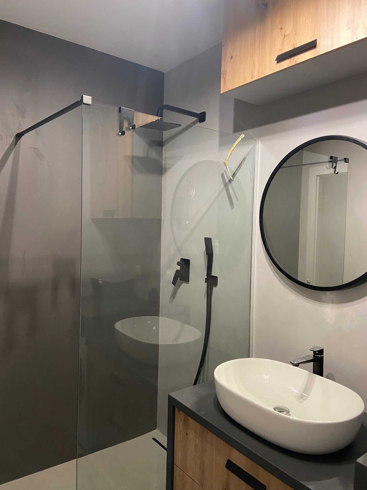 Nowoczesna łazienka z kabiną typu walk-in, owalną umywalką nablatową i okrągłym lustrem w czarnej ramie. Minimalistyczny design z elementami drewna i szarości.