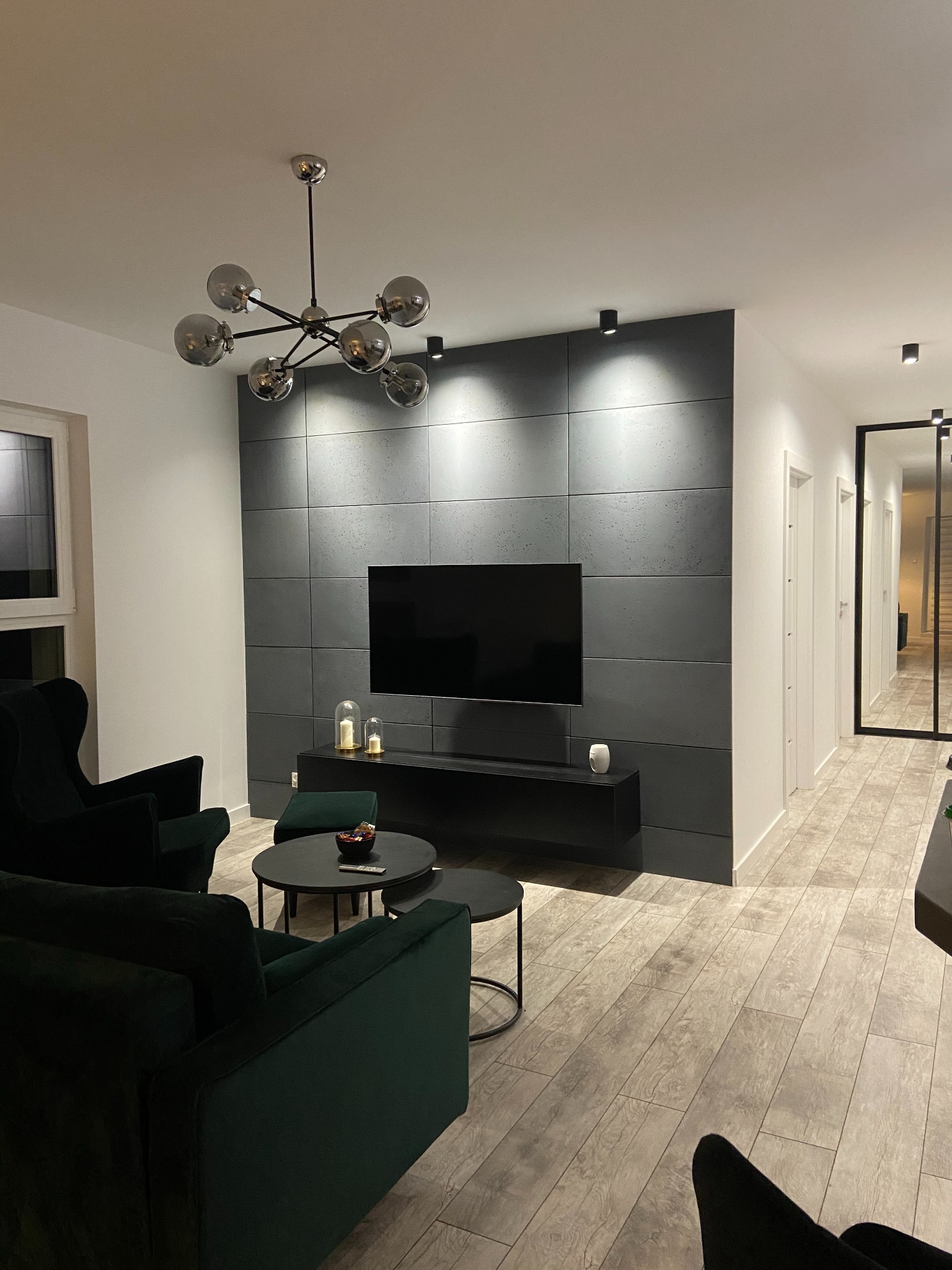 Stylowy salon z szarymi panelami ściennymi, czarnym telewizorem i wiszącą lampą. Wnętrze dopełniają zielone fotele i drewnopodobna podłoga.
