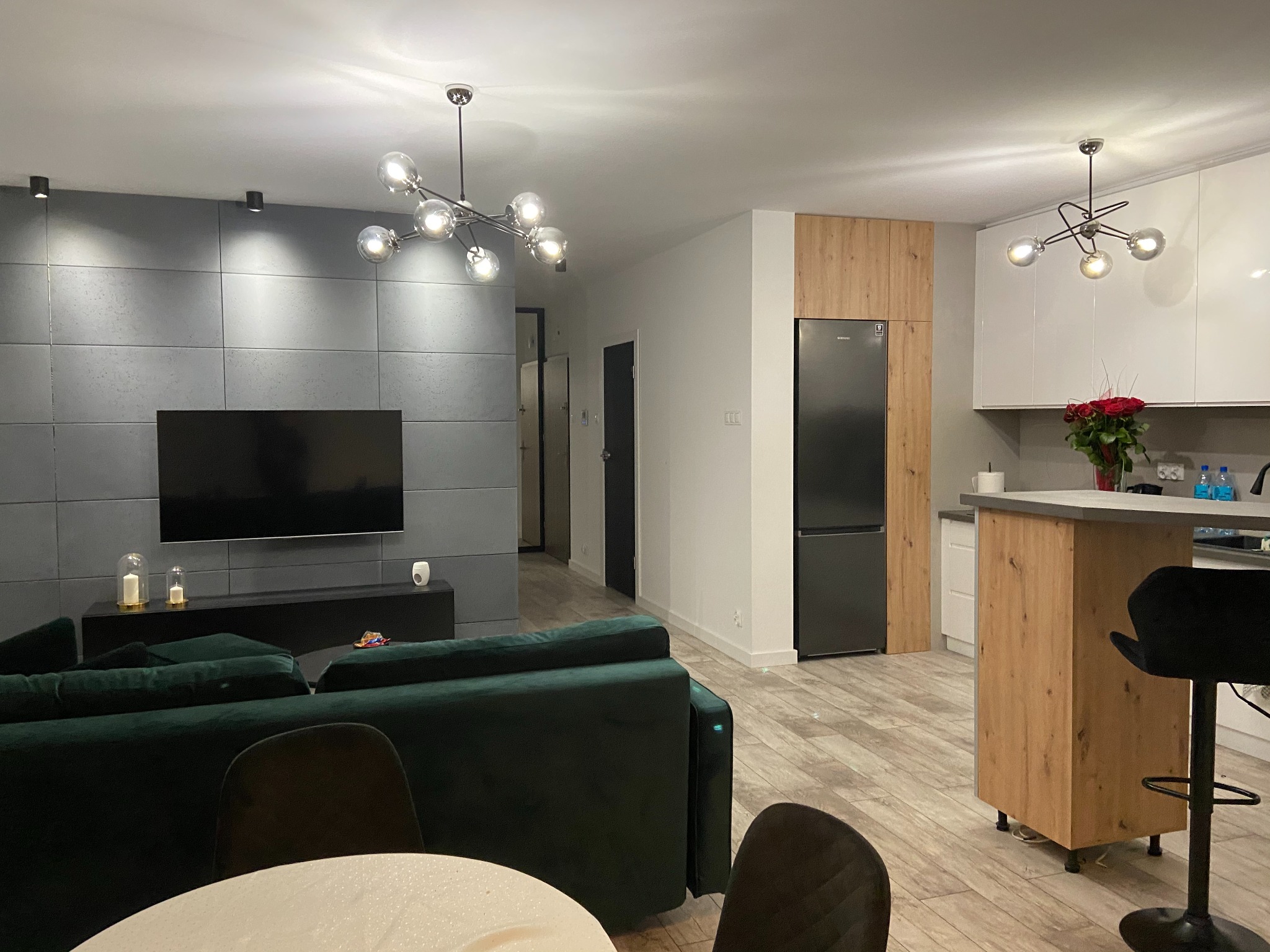 Nowoczesne wnętrze: szara ściana z TV, zielona sofa, drewniane akcenty w kuchni i stylowe lampy wiszące. Parkiet w odcieniach beżu dopełnia aranżacji.