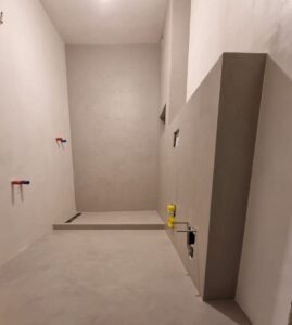 Surowe wnętrze łazienki w odcieniach szarości, z widocznymi instalacjami hydraulicznymi i elektrycznymi przed wykończeniem, ukazujące etap przygotowania do dalszych prac.