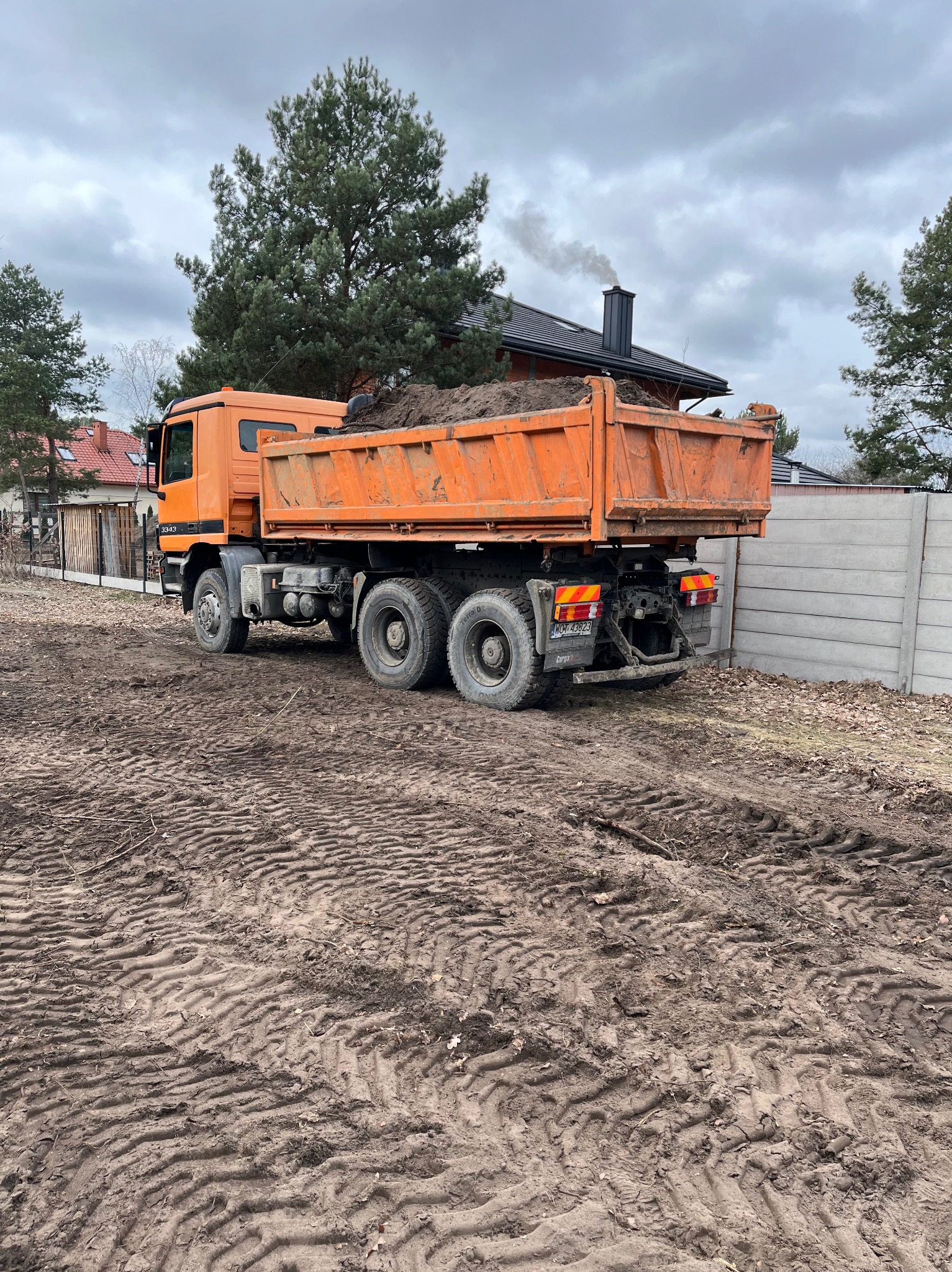 Pomarańczowa wywrotka z ziemią na placu budowy, widoczne ślady opon na ziemi, w tle dom z kominem, z którego wydobywa się dym, pochmurne niebo.