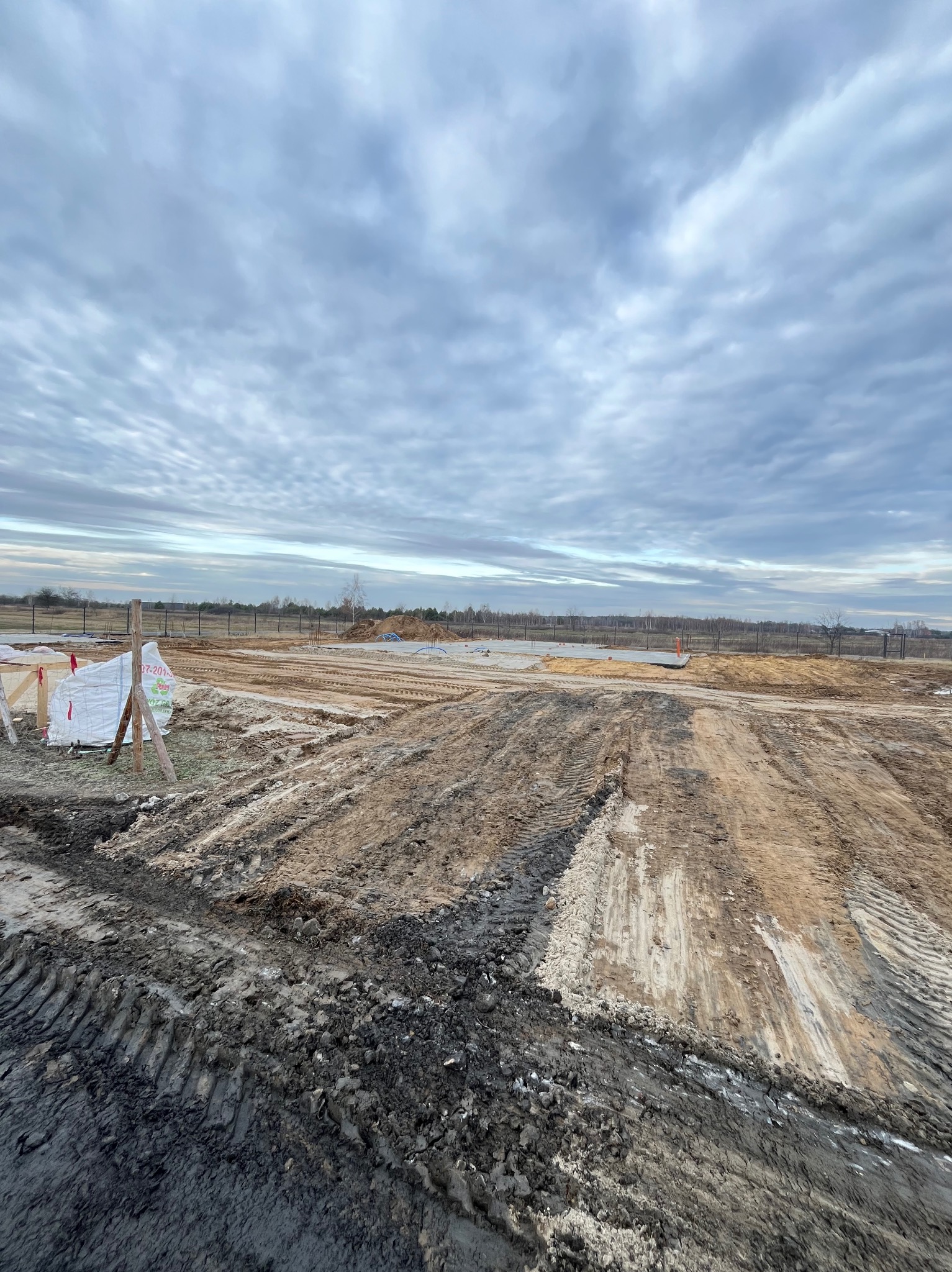 Przygotowany teren budowlany z widocznymi śladami opon, drewnianymi szalunkami i fundamentami w tle, pod pochmurnym niebem.