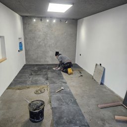 Dziubol Firma Budowlana Jerzy Stokłosa - Glazurnik układa ciemne płytki w pomieszczeniu z szarą ścianą i surowym stropem. Widoczny klej, narzędzia i wiadro z zaprawą. Praca w toku, oświetlenie sufitowe.