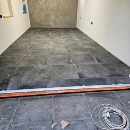 Dziubol Firma Budowlana Jerzy Stokłosa - Wnętrze w trakcie wykańczania: ułożona podłoga z ciemnych, kwadratowych płytek. Listwa progowa z pomarańczowym akcentem. Na podłodze kabel i fragment kartonu.