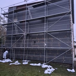 Dziubol Firma Budowlana Jerzy Stokłosa - Dom w trakcie ocieplania styropianem, widoczne rusztowania i resztki materiałów na trawie. Budynek z ciemną elewacją w trakcie prac ociepleniowych.