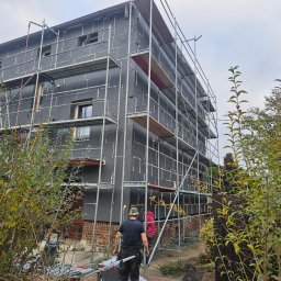 Dziubol Firma Budowlana Jerzy Stokłosa - Dom w trakcie ocieplania elewacji, widoczne rusztowania i płyty izolacyjne. Pracownik na chodniku, fragment ogrodu z krzewami. Pochmurne niebo.