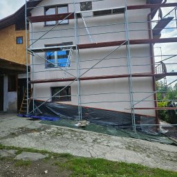 Dziubol Firma Budowlana Jerzy Stokłosa - Dom w trakcie ocieplania elewacji, widoczne rusztowania, zabezpieczona folią podstawa budynku, fragment elewacji z oknami i drewnianym wykończeniem.
