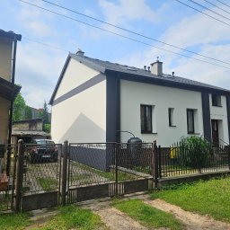 Dziubol Firma Budowlana Jerzy Stokłosa - Odnowiona fasada dwukondygnacyjnego domu z ciemnymi akcentami, widoczna zza metalowego ogrodzenia i zieleni. Uwagę zwraca kontrast bieli ścian i grafitowych elementów wykończenia.