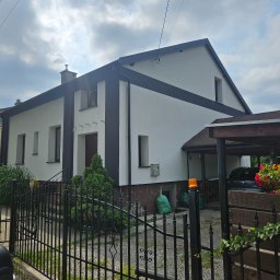 Dziubol Firma Budowlana Jerzy Stokłosa - Dwukondygnacyjny dom z białą elewacją i czarnymi akcentami, widoczny zza czarnego, kutego ogrodzenia. Zielone worki i roślinność przed budynkiem. Pochmurne niebo w tle.