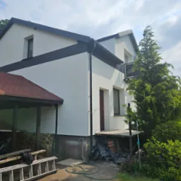 Elewacja domu po malowaniu z czarnym pasem, widoczne narzędzia i samochód w wiacie. Dom otoczony zielenią, jasne niebo z chmurami w tle.