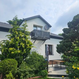 Dziubol Firma Budowlana Jerzy Stokłosa - Biały dom z ciemną dachówką i ozdobnym balkonem. Widoczny ogród z żółtymi różami i licznymi drzewami iglastymi. Na ścianie satelita.