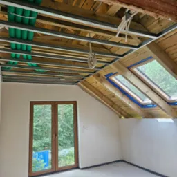 Poddasze w trakcie adaptacji: widoczne okna dachowe, instalacja pod sufitem z rurami wentylacyjnymi i profile do płyt gipsowych. Ściany przygotowane do dalszych prac wykończeniowych.