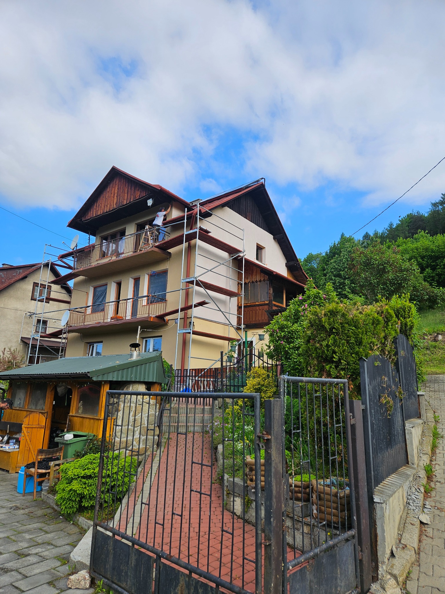 Dwupiętrowy dom w trakcie prac elewacyjnych, z rusztowaniami i pracownikiem przy dachu, na tle błękitnego nieba z chmurami.