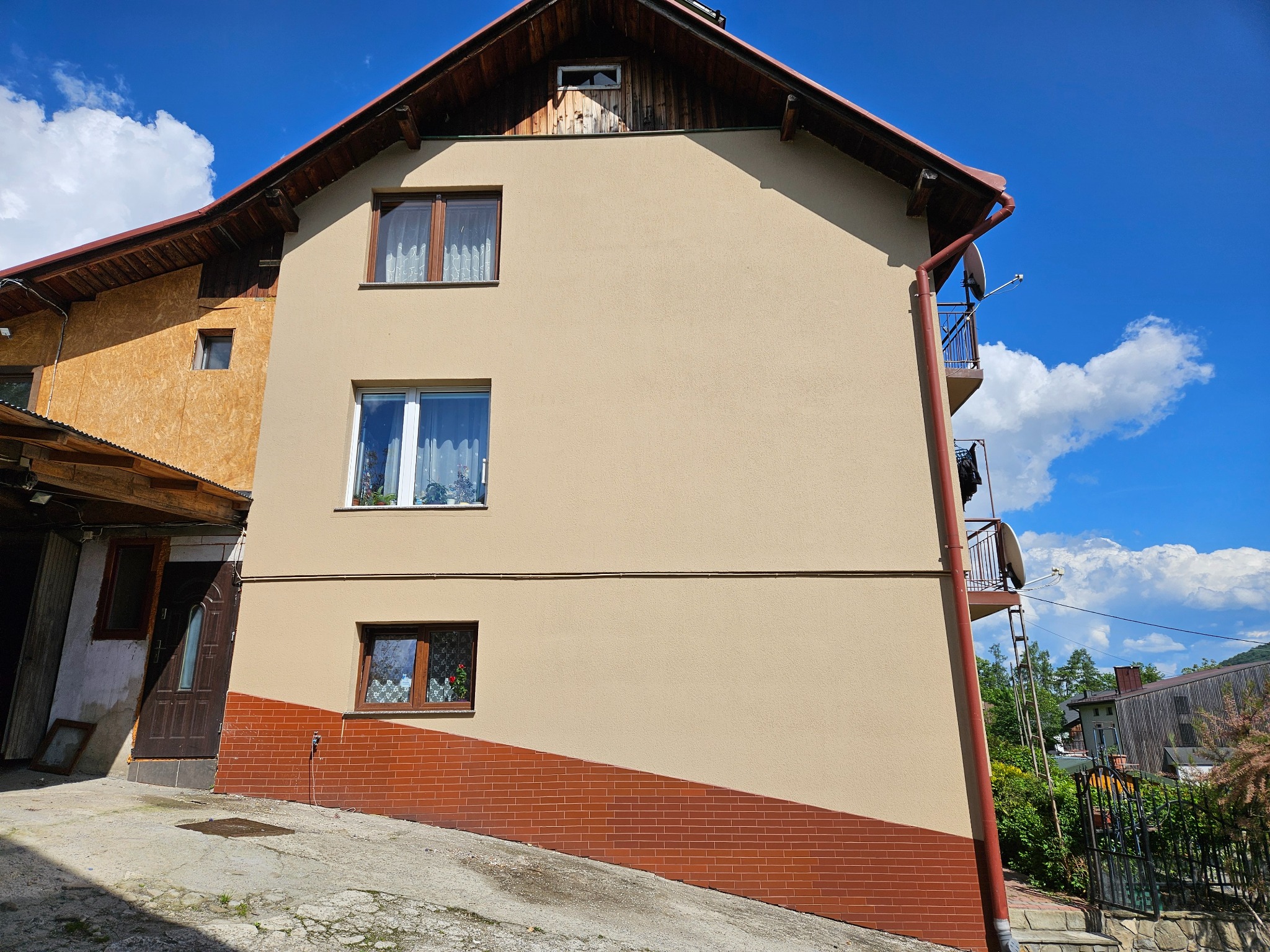 Elewacja domu z oknami i balkonem, wykończona w beżowym kolorze z ceglanym cokołem. Widoczne detale architektoniczne i fragment nieba z chmurami.
