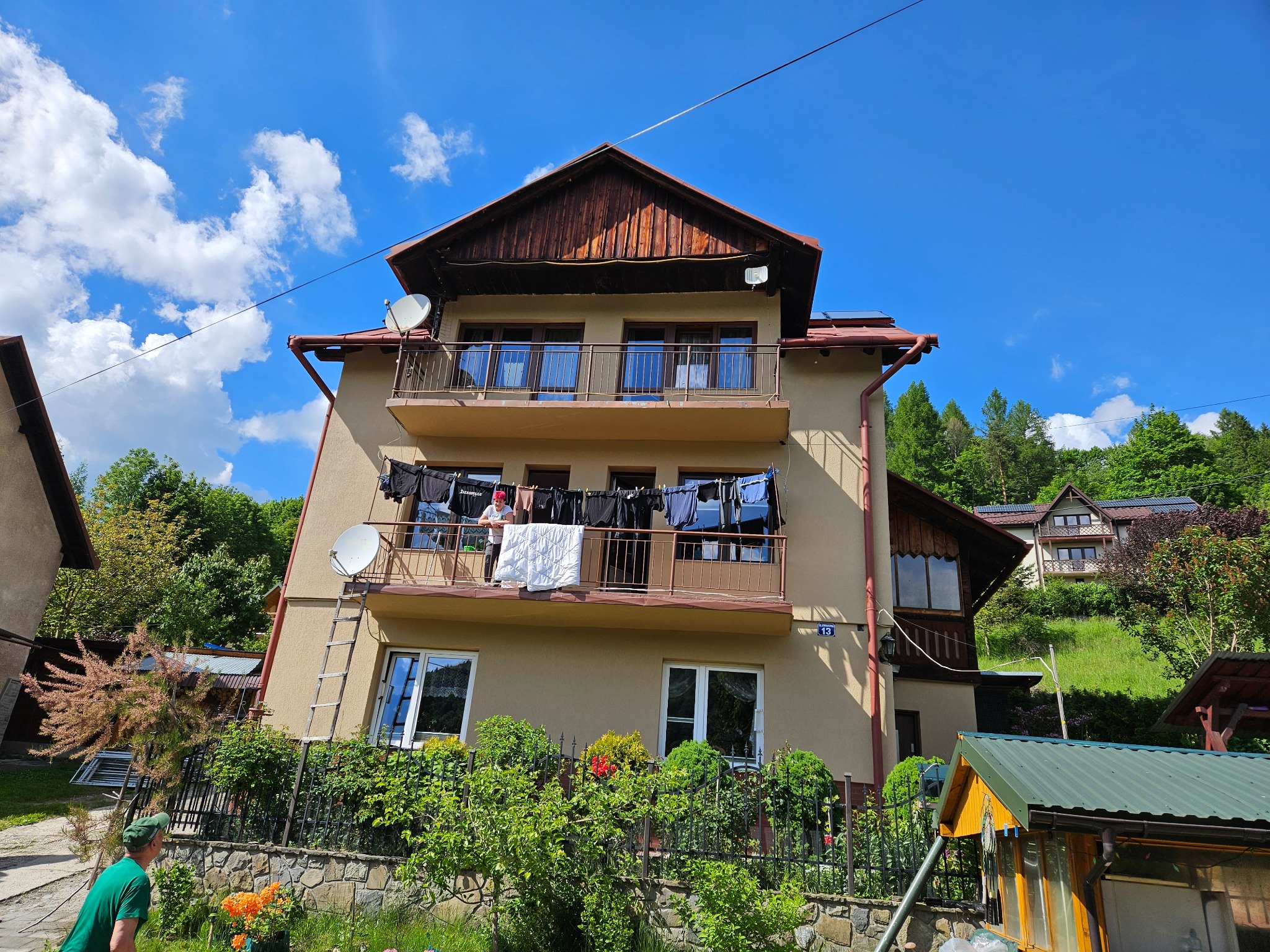 Trzykondygnacyjny dom z beżową elewacją i drewnianymi elementami. Na balkonie suszące się pranie. Widoczne anteny satelitarne i zielone otoczenie.