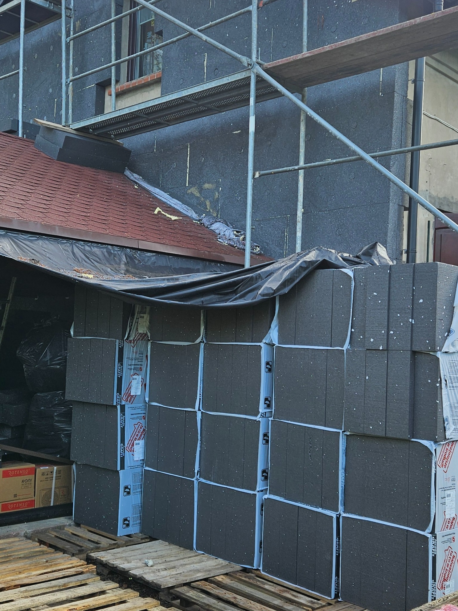 Ocieplanie budynku grafitowym styropianem, przygotowane pakiety na paletach, rusztowanie przy ścianie. Etap prac ociepleniowych.