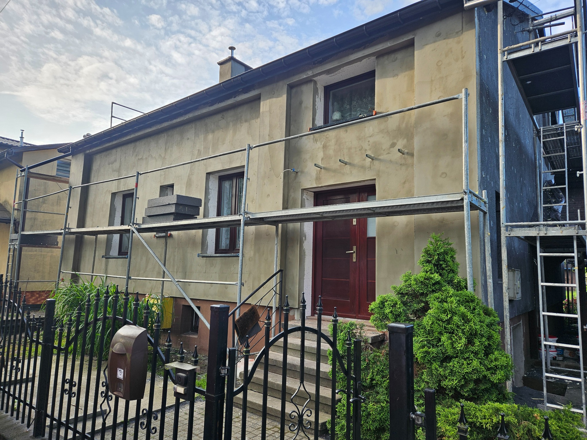 Dom w trakcie prac elewacyjnych: rusztowania, surowy tynk, częściowo ocieplona ściana boczna. Widok zza ozdobnego, metalowego ogrodzenia.