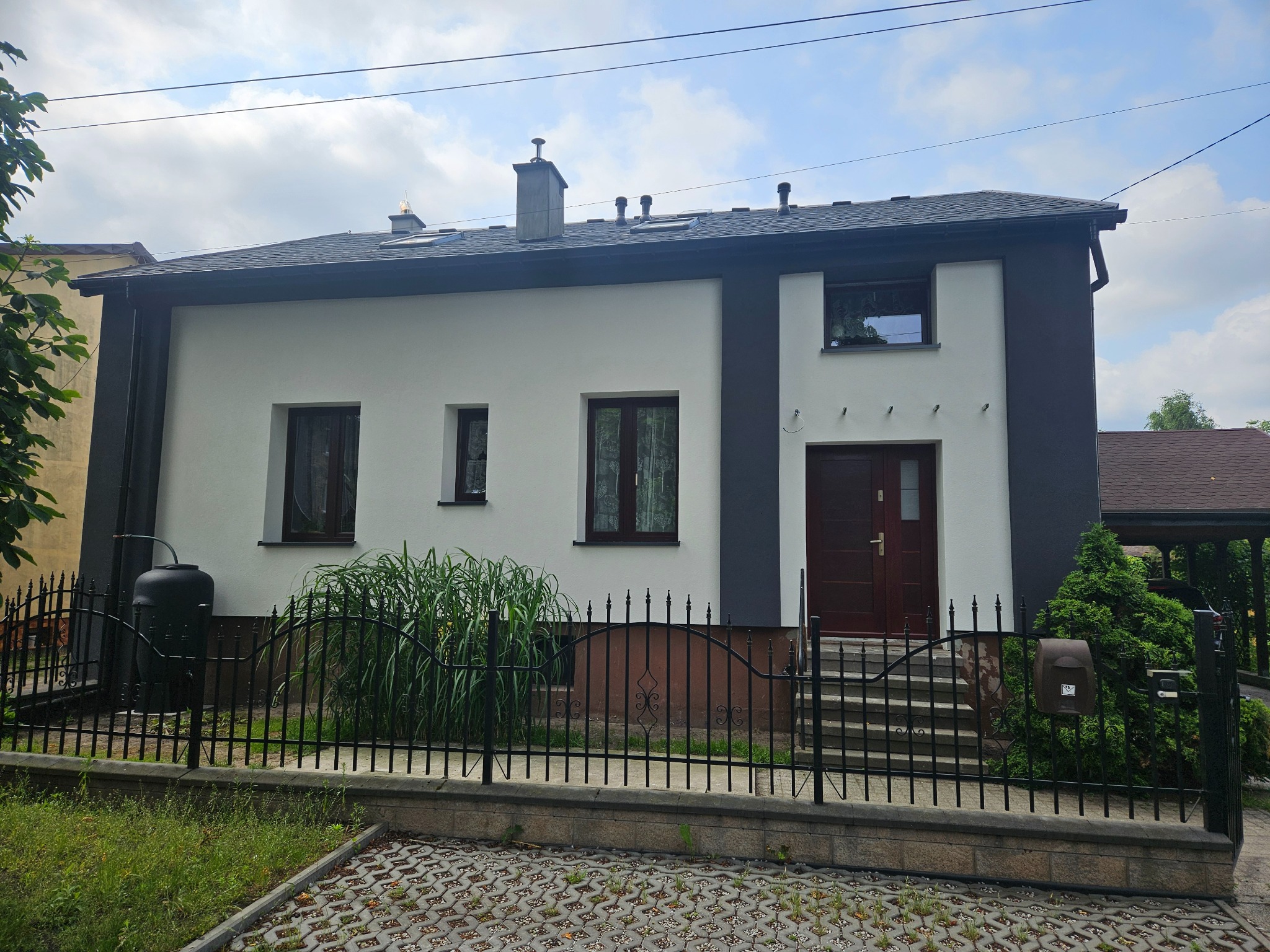 Dwukondygnacyjny dom z białą elewacją i grafitowymi akcentami przy oknach i drzwiach, otoczony czarnym ogrodzeniem i zielenią. Widoczny zbiornik na deszczówkę.
