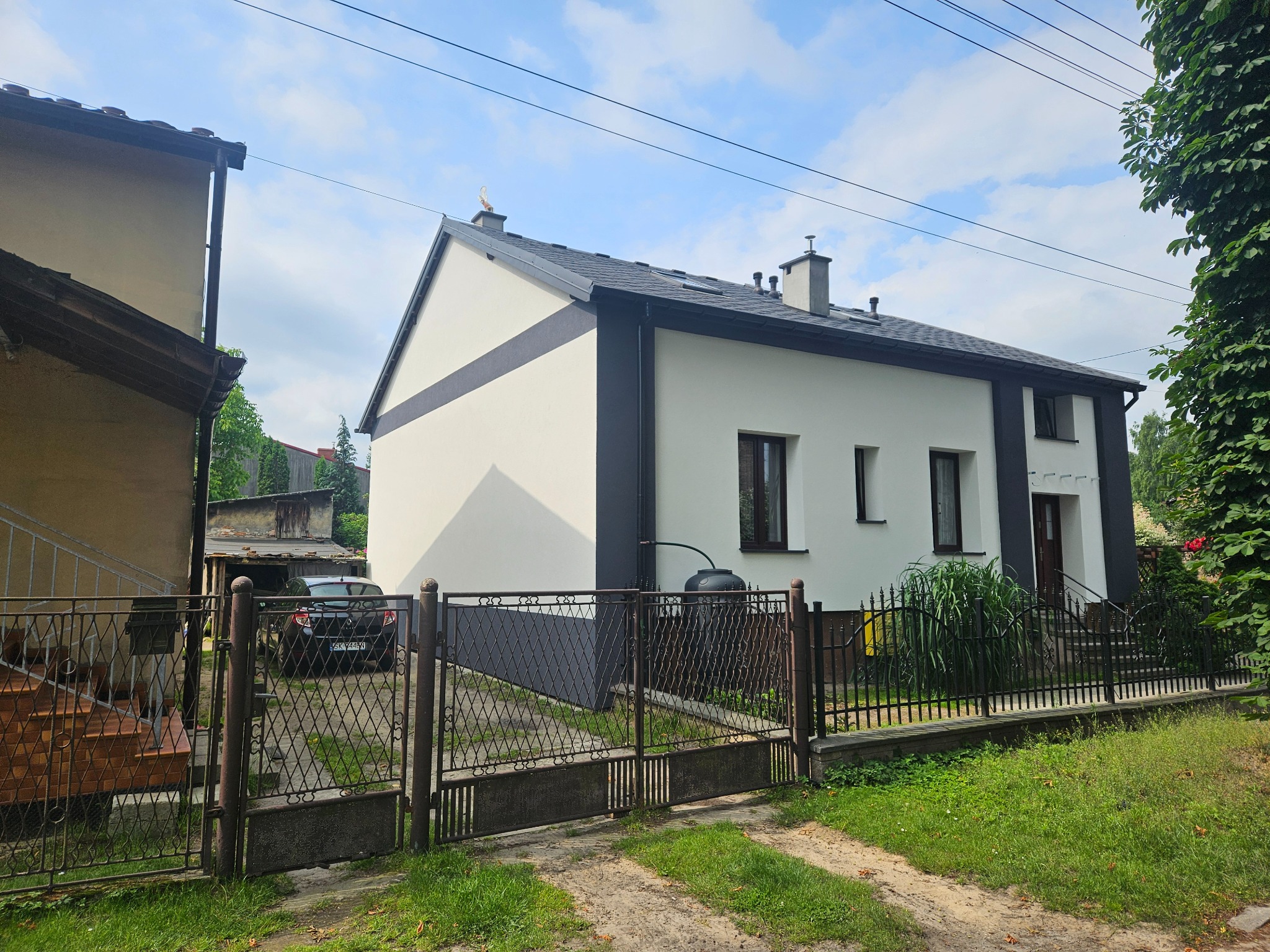 Odnowiona fasada dwukondygnacyjnego domu z ciemnymi akcentami, widoczna zza metalowego ogrodzenia i zieleni. Uwagę zwraca kontrast bieli ścian i grafitowych elementów wykończenia.