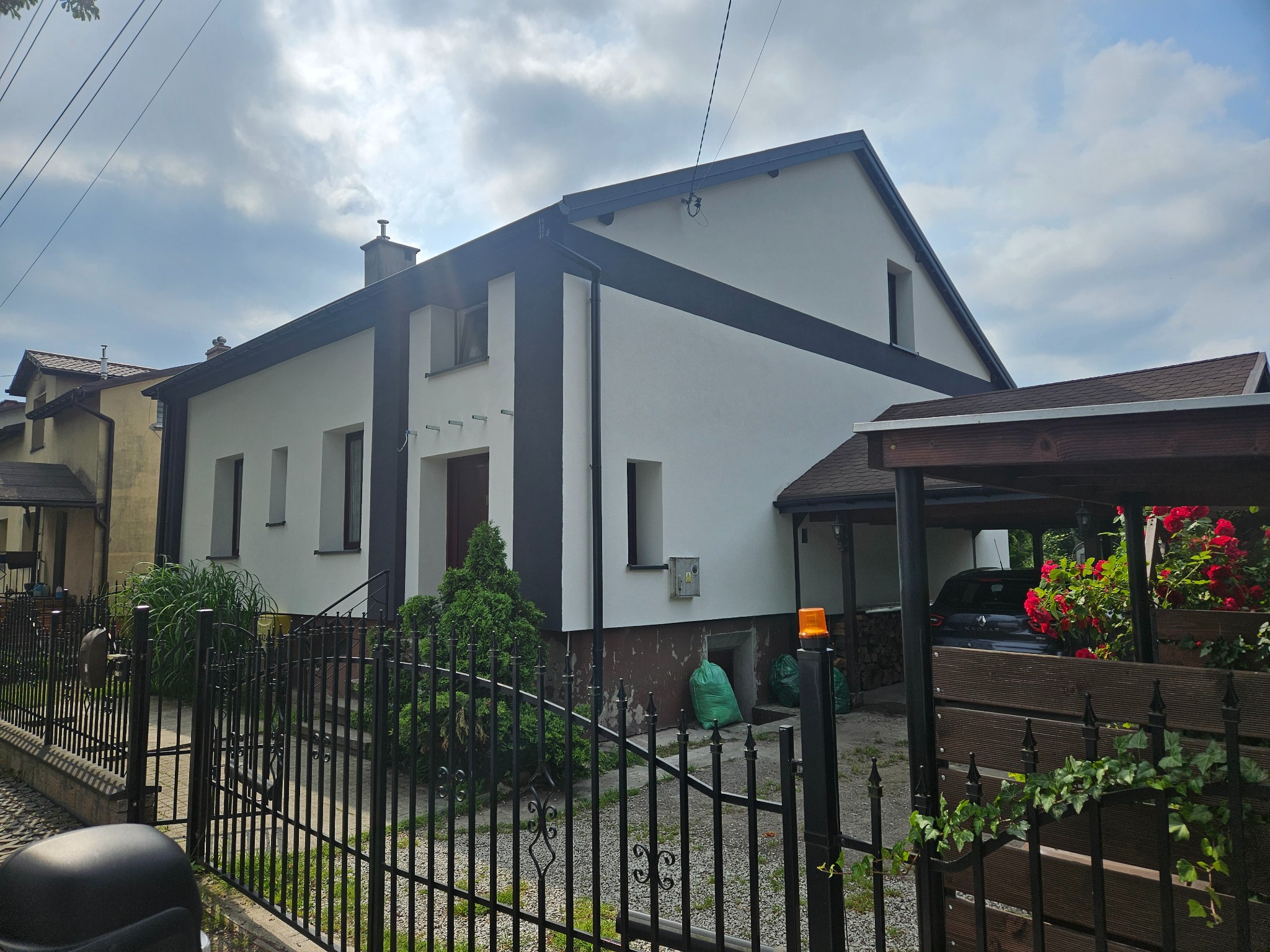 Dwukondygnacyjny dom z białą elewacją i czarnymi akcentami, widoczny zza czarnego, kutego ogrodzenia. Zielone worki i roślinność przed budynkiem. Pochmurne niebo w tle.