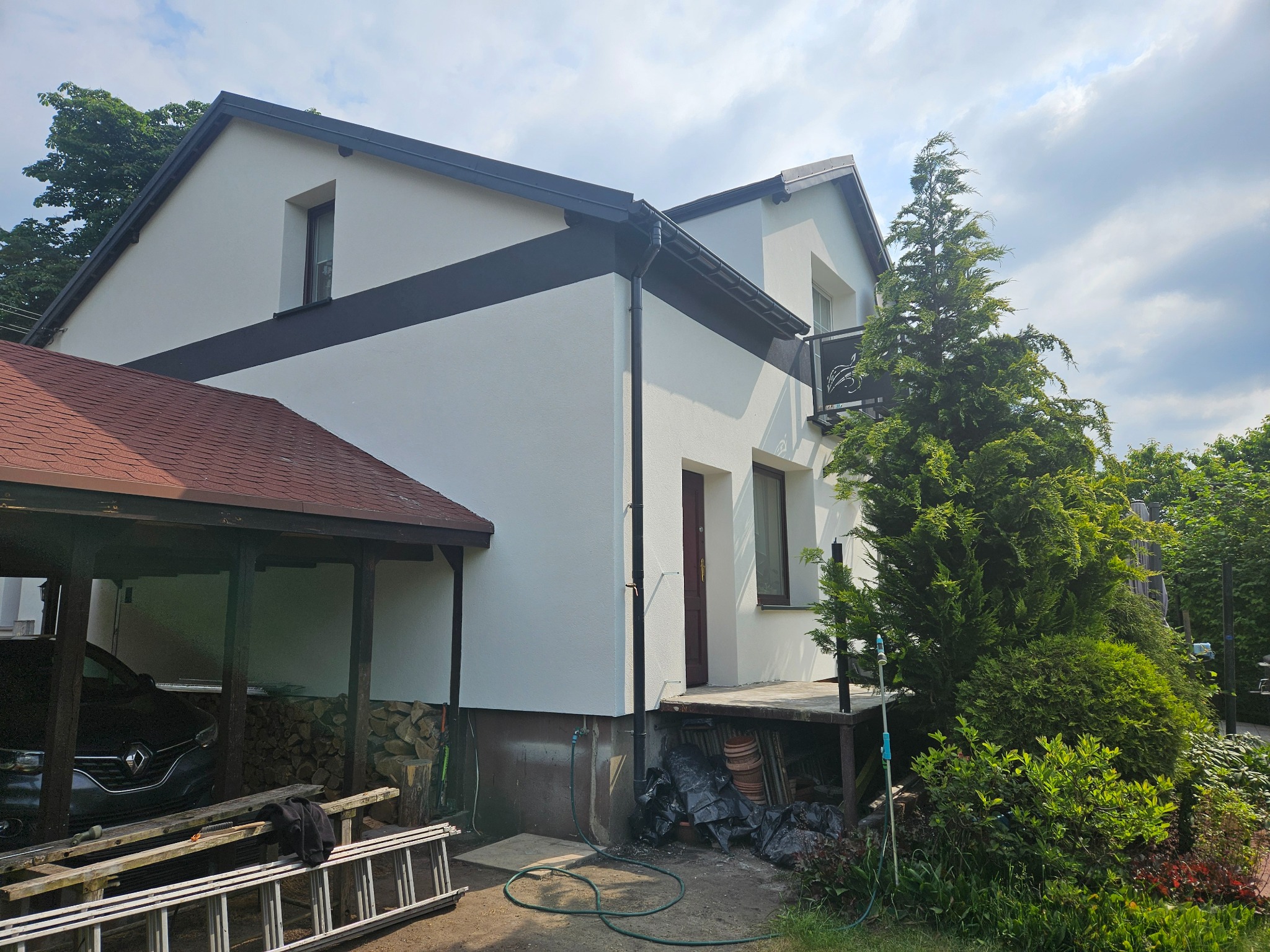 Elewacja domu po malowaniu z czarnym pasem, widoczne narzędzia i samochód w wiacie. Dom otoczony zielenią, jasne niebo z chmurami w tle.