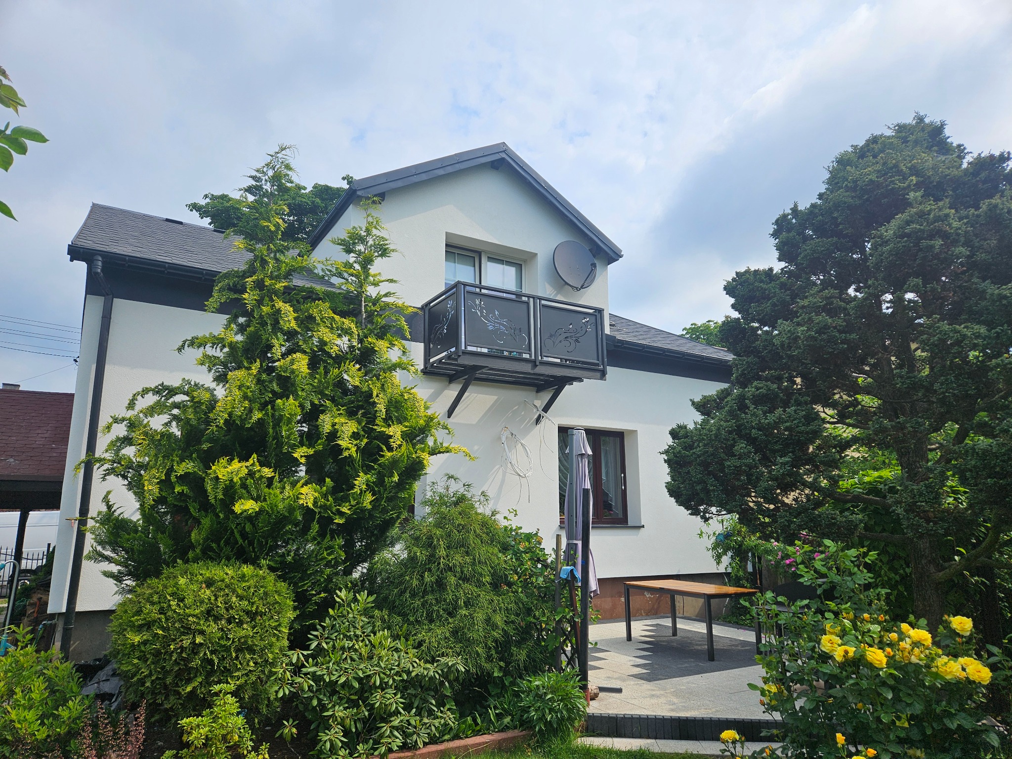 Biały dom z ciemną dachówką i ozdobnym balkonem. Widoczny ogród z żółtymi różami i licznymi drzewami iglastymi. Na ścianie satelita.