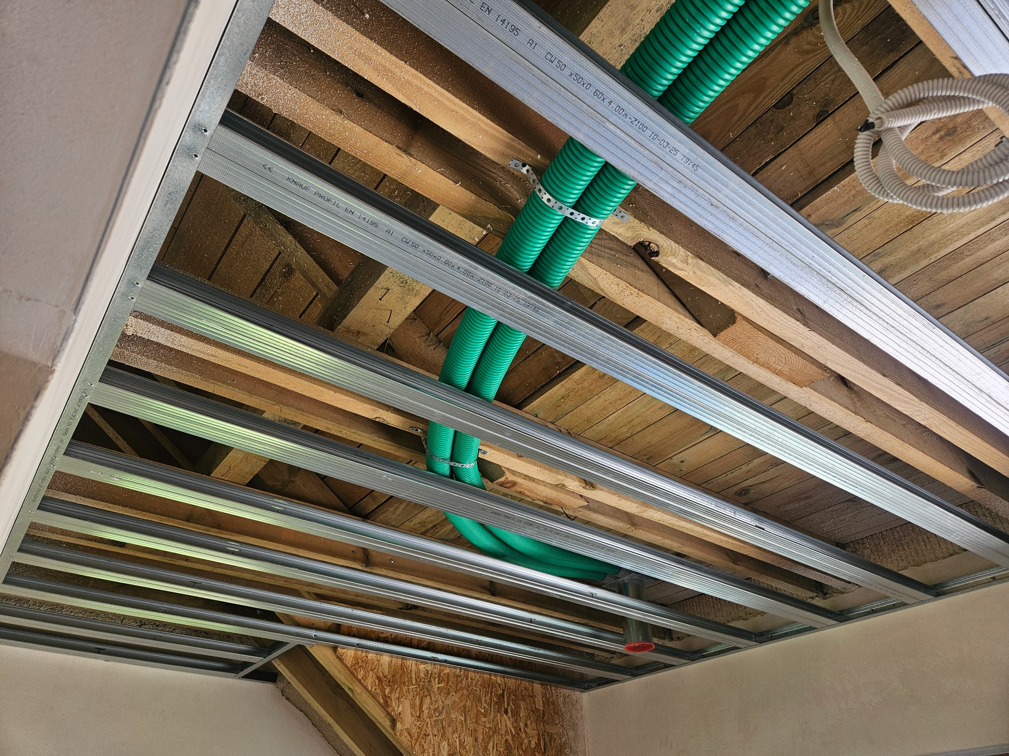 Montaż metalowego stelaża sufitu podwieszanego z widocznymi zielonymi rurami instalacji elektrycznej i drewnianą konstrukcją nośną. Kątowe ujęcie, surowy stan.