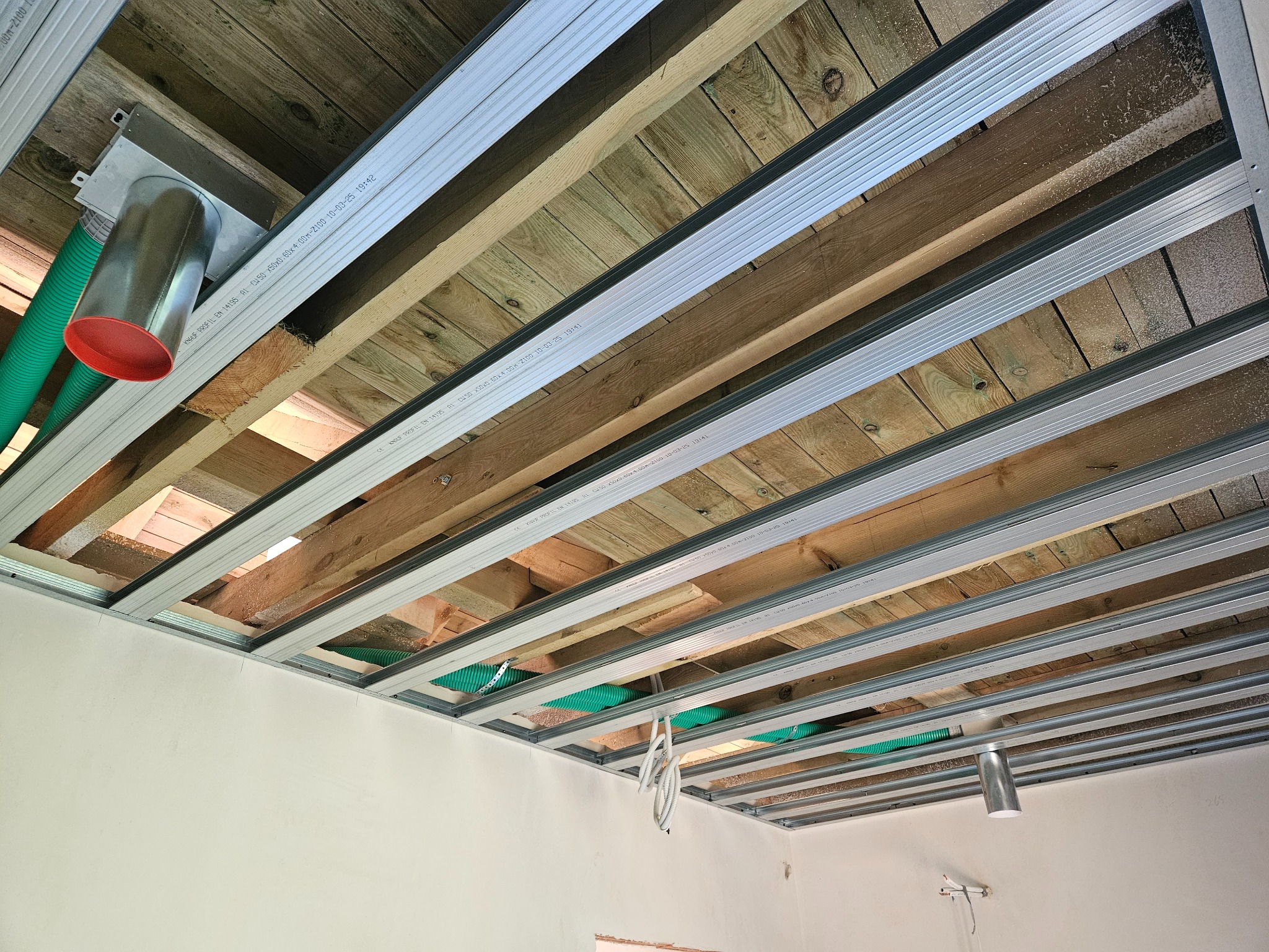 Montaż metalowej konstrukcji sufitu podwieszanego z widoczną instalacją wentylacyjną i drewnianą konstrukcją stropu. Widok z dołu, surowy stan.