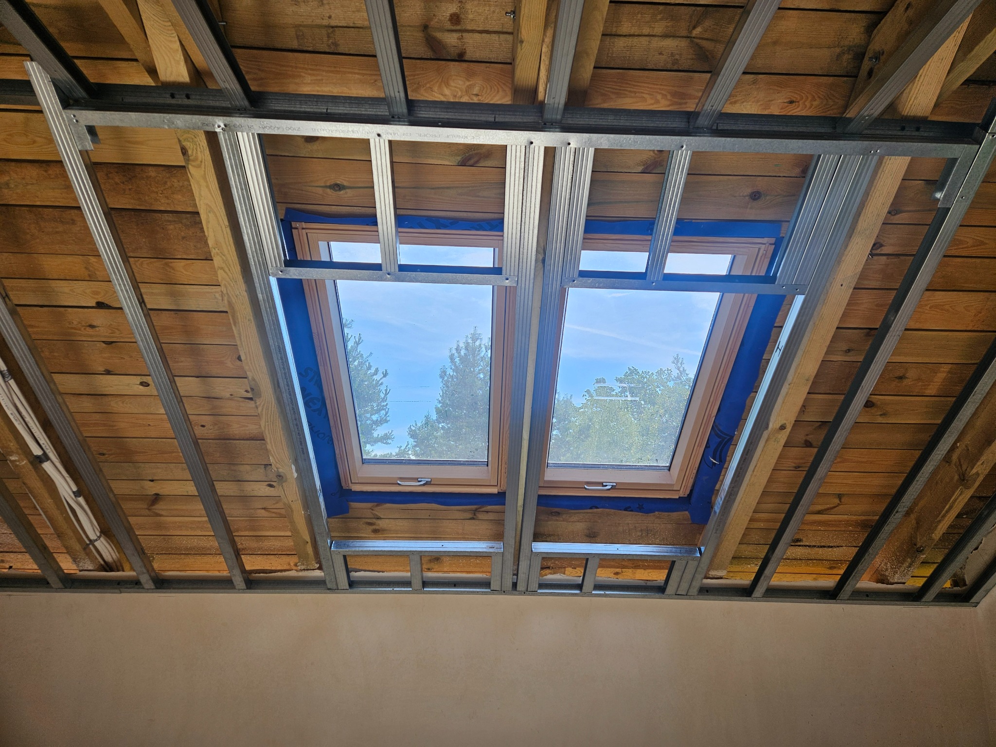 Montaż sufitu podwieszanego z płyt karton-gips na drewnianym poddaszu z oknami dachowymi. Widoczne profile stalowe i drewniana konstrukcja dachu.